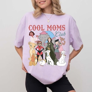 May include: A light purple t-shirt with the text "COOL MOMS Club" in red and blue. The shirt features cartoon illustrations of various characters, including a lion, dog, and cat. The shirt is made of a soft material.
