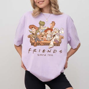 May include: A light purple t-shirt featuring a graphic of Toy Story characters sitting on a couch. The word "FRIENDS" is printed in brown, with "SINCE 1995" below it. The shirt has short sleeves.