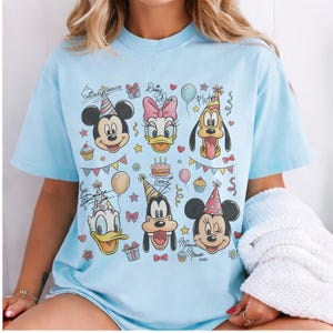 Puede incluir: Camiseta azul claro con los personajes de dibujos animados Mickey Mouse, Minnie Mouse, Donald Duck, Daisy Duck, Goofy y Pluto. El diseño incluye elementos de fiesta de cumpleaños como globos, cupcakes y confeti.