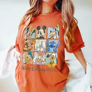 Puede incluir: Camiseta naranja quemado con una cuadr&iacute;cula de personajes de Disney, incluyendo Mickey Mouse, Minnie Mouse, Goofy, Chip y Chop, Stitch, Donald Duck y Daisy Duck. Las palabras "Walt Disney World" est&aacute;n impresas en cursiva.