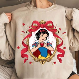 Pode incluir: Camisola bege com uma ilustração emoldurada da Branca de Neve segurando uma maçã. A ilustração é cercada por uma fita vermelha e detalhes florais. O design inclui uma moldura dourada e um laço vermelho.