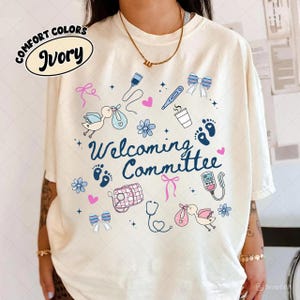 Peut inclure: T-shirt ivoire avec un motif circulaire comprenant des illustrations liées aux bébés et les mots "Welcoming Committee". Le motif comprend des cigognes, des empreintes de pas, des nœuds et des outils médicaux. Le t-shirt est de la marque Comfort Colors.