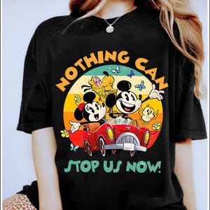 May include: Black t-shirt featuring a colorful graphic of Mickey and Minnie Mouse in a red car, with the text "NOTHING CAN STOP US NOW!". The design includes butterflies, flowers, and other cartoon characters. The shirt has a crew neck.