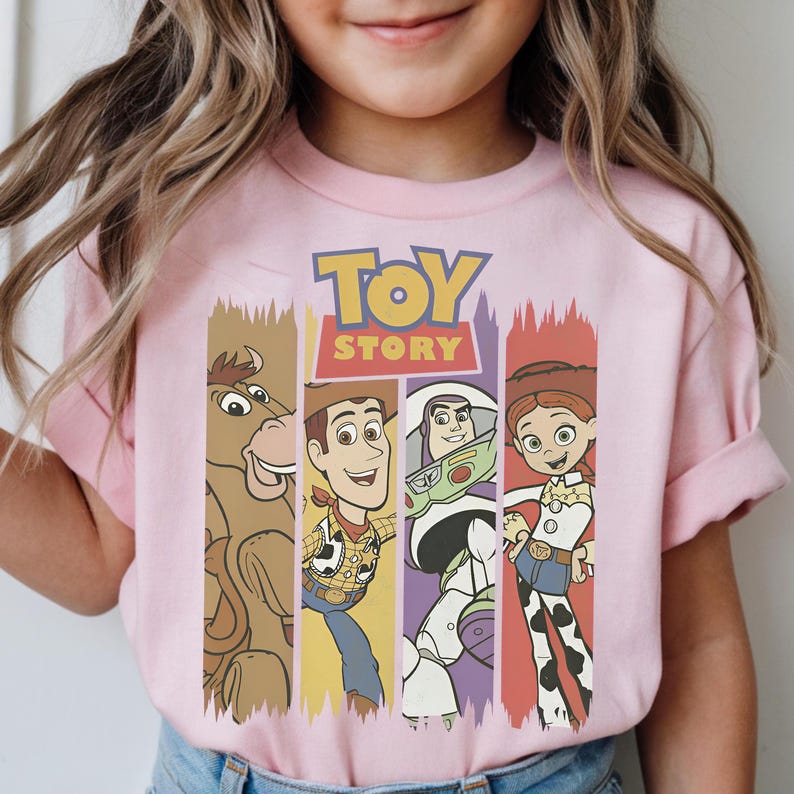 May include: Pink t-shirt featuring the Toy Story logo and characters. The design includes images of Bullseye, Woody, Buzz Lightyear, and Jessie, each within a vertical stripe. The shirt is made of a soft material.