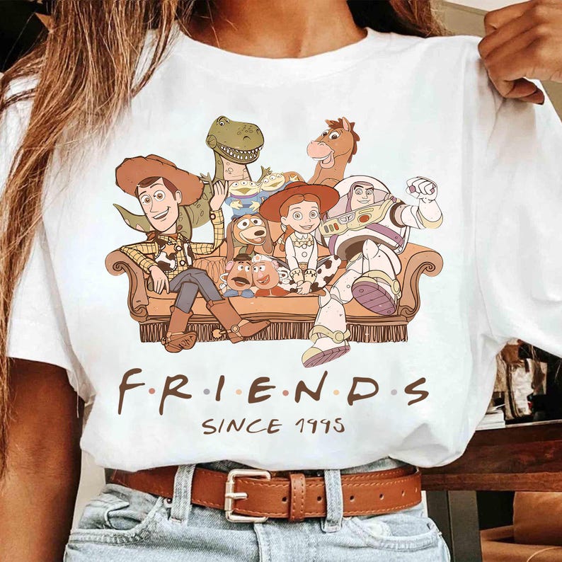 May include: White t-shirt featuring a graphic of characters from the animated film Toy Story, including Woody, Buzz Lightyear, and others, seated on a brown couch. The text "FRIENDS SINCE 1995" is printed below the graphic.