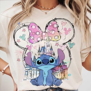 May include: Cream-colored t-shirt featuring a cartoon illustration of Stitch from Lilo & Stitch. The design includes a pink polka-dot bow, a castle, and Mickey Mouse-shaped balloons. The artwork is outlined in black.