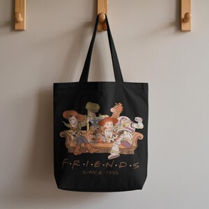 May include: Black tote bag featuring a colorful illustration of characters from the Toy Story movie franchise, seated on a brown couch. The word "FRIENDS" is printed in white, with "SINCE 1995" below.