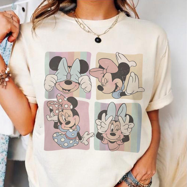 May include: Cream-colored t-shirt featuring a four-panel design of Minnie Mouse. Each panel showcases a different pose of Minnie, with pastel-colored backgrounds. Minnie is wearing bows and dresses in various colors.