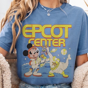 May include: A light blue t-shirt featuring the words "EPCOT CENTER" in yellow and red block letters. The graphic depicts Mickey Mouse and Figment in space suits on a planet with planets and stars in the background.