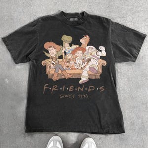 May include: Dark gray t-shirt featuring a graphic of characters from the animated film Toy Story, seated on a couch. The word "FRIENDS" is printed below the image, with "SINCE 1995" underneath.