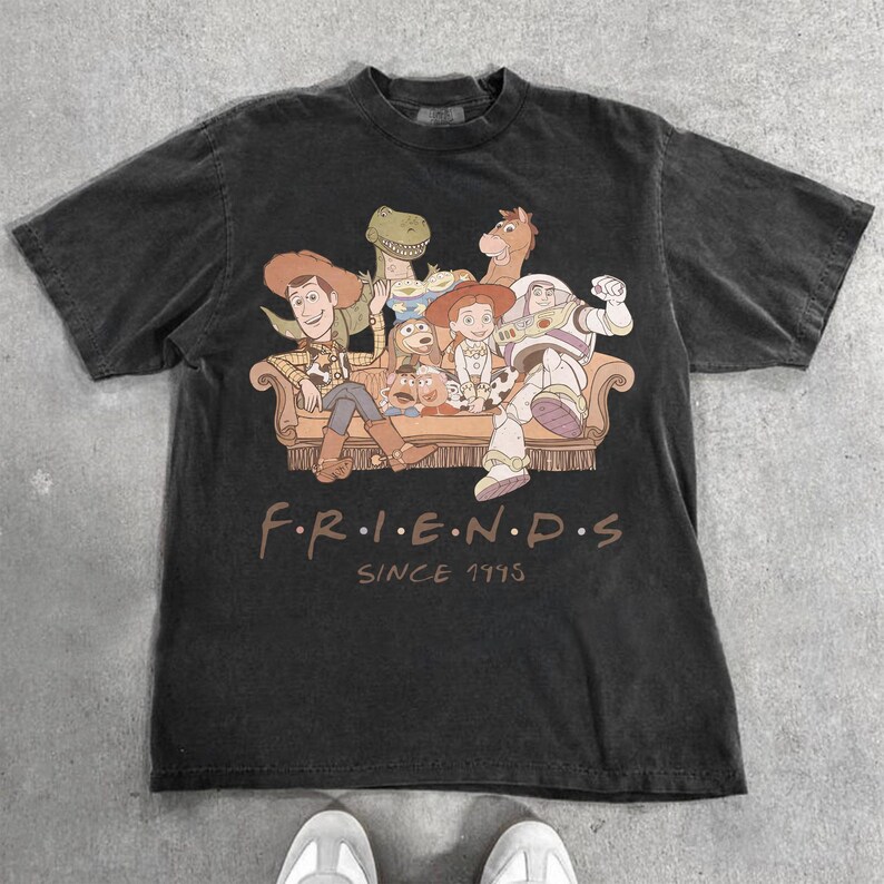 May include: Dark gray t-shirt featuring a graphic of characters from the animated film Toy Story, seated on a couch. The word "FRIENDS" is printed below the image, with "SINCE 1995" underneath.
