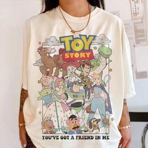 Pu&ograve; includere: T-shirt color crema con una grafica colorata dei personaggi del film d'animazione "Toy Story". Il design include il logo del film e la frase "You've Got a Friend in Me".