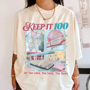 May include: Cream-colored t-shirt featuring a colorful graphic with the text "KEEP IT 100." The graphic includes images of a geometric structure, a monorail, a boat, and seagulls. The phrase "On The Land. The Seas. The Skies" is also printed on the shirt.