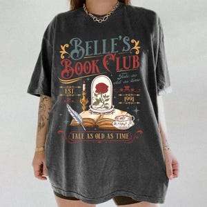 May include: Dark gray t-shirt featuring the text "Belle's Book Club" with a rose in a glass dome, a teacup, and a feather quill. The design includes the words "Tale as old as time" and the year 1991.