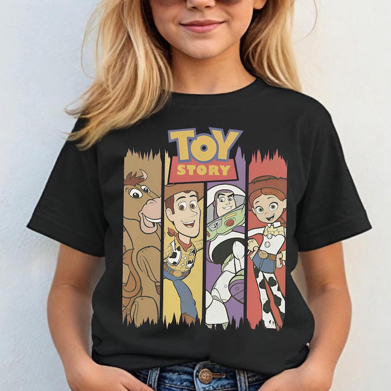 May include: Black t-shirt featuring the Toy Story logo and characters. The design includes images of Bullseye, Woody, Buzz Lightyear, and Jessie, each in a vertical panel with a different color background. The shirt is worn by a person.