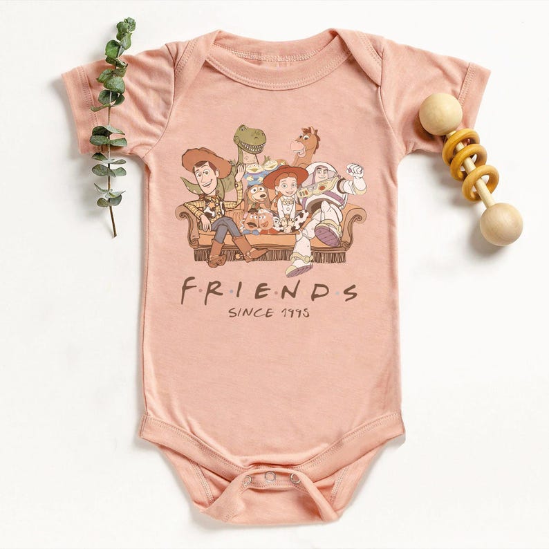 May include: A light pink baby onesie featuring a graphic of characters from the Toy Story movie franchise sitting on a couch. The text "FRIENDS SINCE 1995" is printed below the graphic. A wooden rattle is on the right.