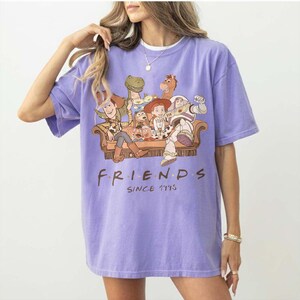 May include: A lilac-colored t-shirt featuring a graphic of characters from the animated film Toy Story, seated on a couch. The word "FRIENDS" is printed below the graphic, with "SINCE 1995" underneath.