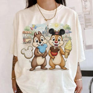 May include: Cream-colored t-shirt featuring a colorful Disney-themed graphic. The graphic depicts Chip and Dale holding treats in front of a park scene with a castle, food cart, and balloons. A Mickey Mouse cookie is also visible.