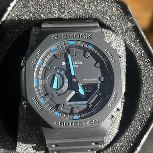 以下が含まれることがあります： 八角形のベゼルを備えたマットブラックのG-SHOCKウォッチ。アナログ針とデジタル表示が特徴です。文字盤のインデックスと針には鮮やかなブルーのアクセントがあり、G-SHOCK、CASIO、WR 20BAR、PROTECTIONの文字が刻まれています。