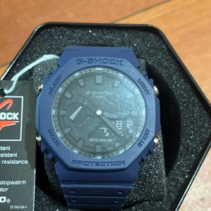 May include: A matte navy blue G-Shock watch with an octagonal bezel, analogue hands, and a digital display window showing the number 22. The watch features silver-tone side buttons labelled Adjust, Light, Mode, and Start, and rests in a black foam insert.