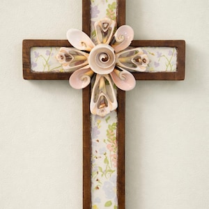 May include: Wooden cross with a dark brown frame, featuring a floral print background. The center of the cross is adorned with a flower made of seashells, with a small gold-colored loop at the top for hanging.