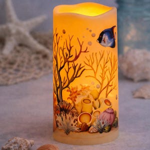May include: A cylindrical candle with a warm glow, featuring a painted underwater scene. The design includes coral, a blue and white fish, and bubbles. Seashells are scattered around the base of the candle, creating a coastal theme.