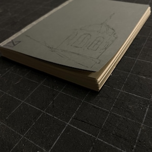 May include: A sketchbook with a gray cover featuring a pencil sketch of a building. The pages are cream-colored, and the book is open, revealing the sketch. The background is a dark surface with a grid pattern.
