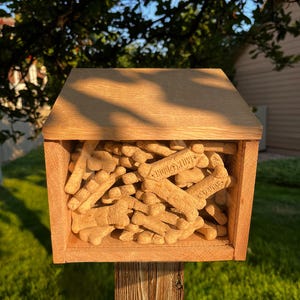 Puede incluir: Una caja de madera llena de galletas para perros, cada una con forma de hueso. Las galletas son de color marrón claro y tienen la inscripción "Milk-Bone". La caja es de madera clara y está montada sobre un poste de madera.