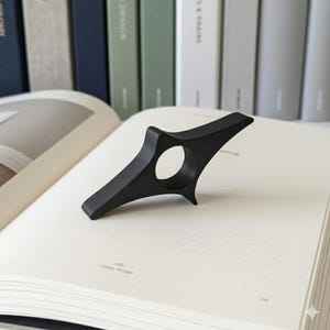 Könnte beinhalten: Ein schwarzer, sternförmiger Buchseitenhalter liegt auf einem offenen Buch. Der Halter hat eine kreisförmige Öffnung für einen Finger und spitze Kanten. Das Buch ist auf einer Seite mit den Worten "Clean design" geöffnet.