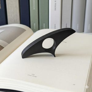 Könnte beinhalten: Ein mattschwarzer Buchseitenhalter liegt auf einem offenen Buch. Der Halter hat ein gebogenes Design mit einer kreisförmigen Öffnung für einen Finger. Das Buch ist auf einer Seite mit den Worten "Clean design" geöffnet.