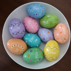 May include: A collection of ten hand-painted Easter eggs in a white bowl. Each egg is a different pastel color and decorated with floral patterns, paw prints, or carrots. Names like "Ben", "Layla", and "Dave" are written on the eggs.