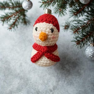 May include: A handmade snowman Christmas ornament. The snowman is crocheted with a white body, a red hat and scarf, a small orange nose, and black eyes. The ornament hangs from a silver metal cap.