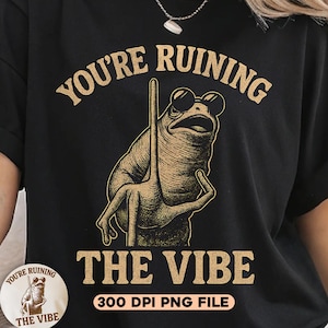 Puede incluir: Camiseta negra con un gráfico de una rana con gafas de sol y sosteniendo un poste. El texto "YOU'RE RUINING THE VIBE" está arqueado sobre la rana, con el mismo texto repetido en un pequeño gráfico circular.