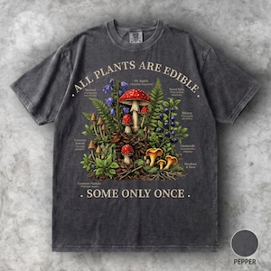 Puede incluir: Una camiseta gris carbón con una ilustración botánica de varios hongos, helechos y bayas. El texto dice ALL PLANTS ARE EDIBLE. SOME ONLY ONCE. El diseño incluye etiquetas para especies como la amanita y el rebozuelo en un estilo vintage.