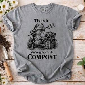 May include: Gray t-shirt with a humorous graphic of a frog holding a pitchfork next to a compost bin. The text on the shirt reads "That's it. You're going in the COMPOST."
