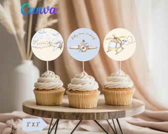 May include: Three cupcakes with white frosting are topped with circular toppers featuring airplane, propeller plane, and helicopter designs. The toppers have text that reads "Baby Jones Incoming!" and "Up Up & Away". The cupcakes sit on a wooden cake stand.
