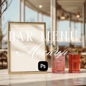 May include: A gold-framed sign with the words "BAR MENU Mockup" is displayed on a marble table. Two glasses of red cocktails are next to the sign. The image has a bright, airy feel, suggesting an outdoor bar setting.
