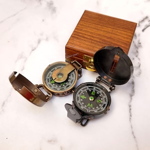 May include: Two antique brass compasses with black dials and green directional indicators are displayed open, next to a wooden box. The compasses have hinged lids and are set against a marble background.