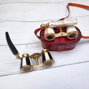 May include: Antique opera glasses with a black handle and gold-colored accents. The binoculars have a black and white striped design. A red leather case with the text "ODDLY CRAFT" is also visible.