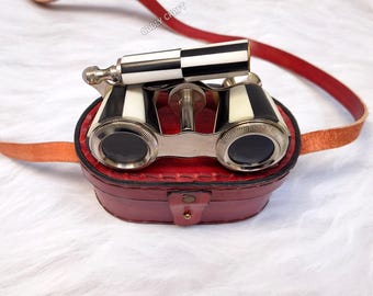 May include: Vintage opera glasses with a black and white striped handle and accents, resting in a red leather case. The case has a matching red leather strap. The text "ODDLY CRAFT" is visible.