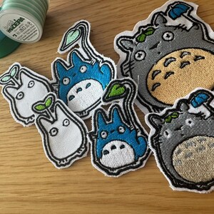 May include: Embroidered patches featuring characters from a popular animated film. The patches include various designs of a large grey creature with a tan belly, and smaller blue and white creatures with leaf accents. A spool of green thread is in the background.