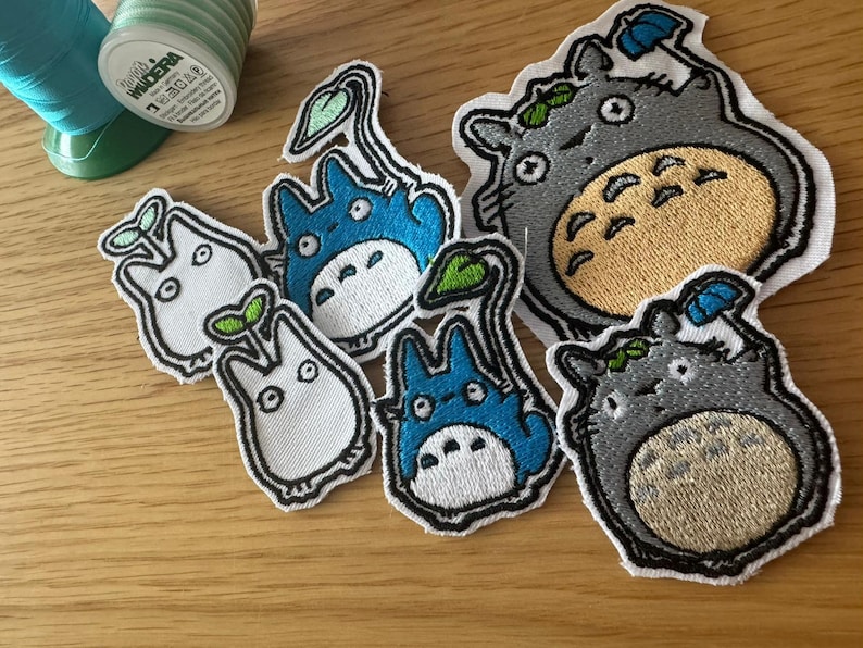 May include: Embroidered patches featuring characters from a popular animated film. The patches include various designs of a large grey creature with a tan belly, and smaller blue and white creatures with leaf accents. A spool of green thread is in the background.