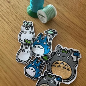 May include: Embroidered patches of cartoon characters in blue, grey, and white, with black outlines. Two spools of thread, one teal and one light green, are also visible. The patches vary in size.