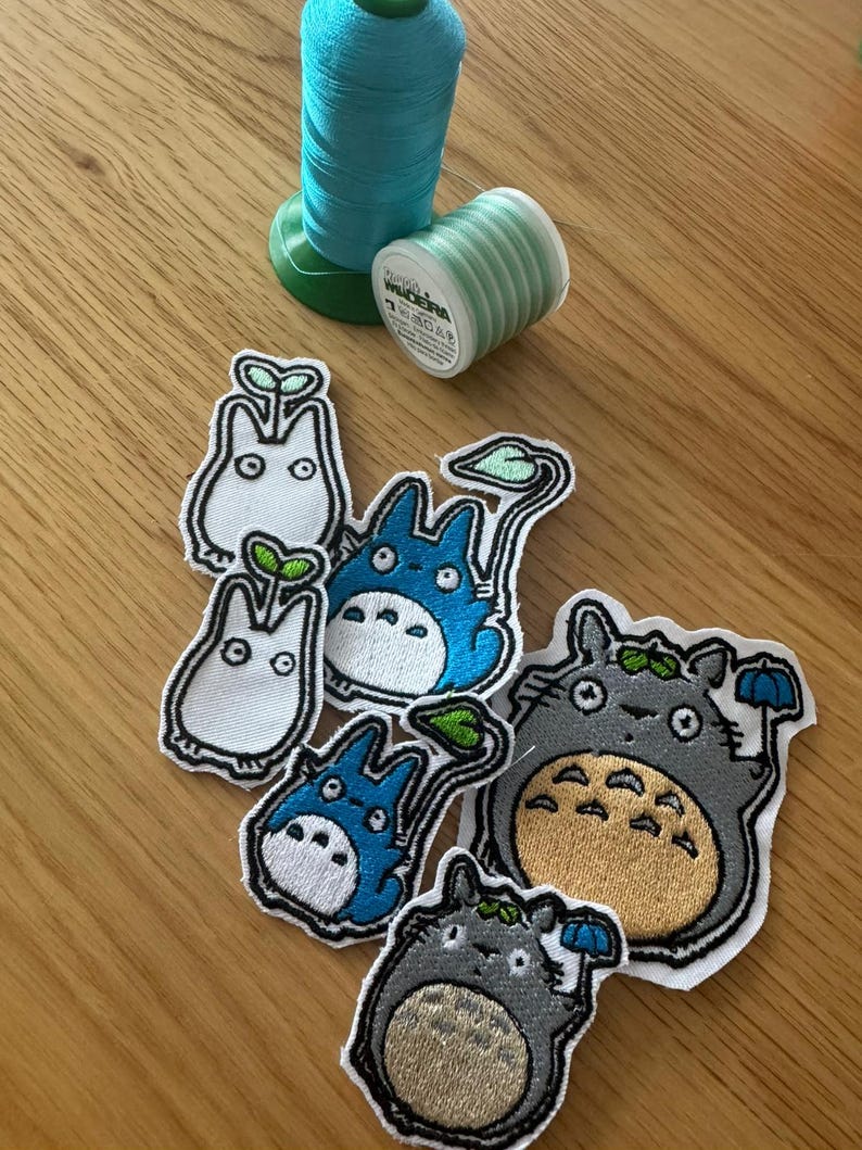 May include: Embroidered patches of cartoon characters in blue, grey, and white, with black outlines. Two spools of thread, one teal and one light green, are also visible. The patches vary in size.