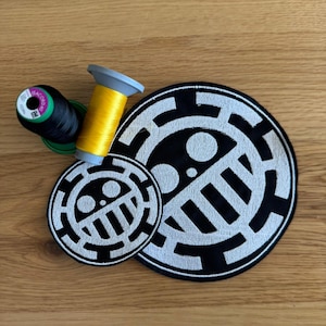 May include: Two black and white embroidered patches with a circular design, and two spools of thread in black and yellow. The patches feature a stylized emblem. The items are arranged on a wooden surface.