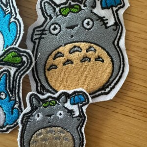 May include: Embroidered patches of the Totoro character from the animated film. The patches are grey with a tan belly, holding a blue umbrella. The patches have a white border and green leaf details.