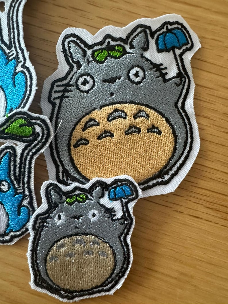 May include: Embroidered patches of the Totoro character from the animated film. The patches are grey with a tan belly, holding a blue umbrella. The patches have a white border and green leaf details.