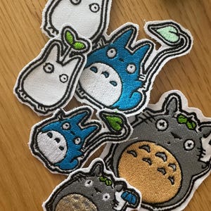May include: A selection of embroidered patches depicting characters from "My Neighbour Totoro." The patches feature white, blue, and grey Totoro designs in various sizes. Each patch has a black outline and is on a white background. The patches are for decorative use.