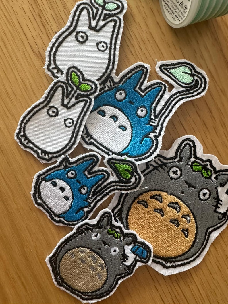 May include: A selection of embroidered patches depicting characters from "My Neighbour Totoro." The patches feature white, blue, and grey Totoro designs in various sizes. Each patch has a black outline and is on a white background. The patches are for decorative use.