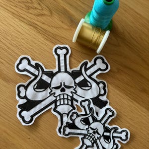 Può includere: Tre toppe bianche e nere con teschio e ossa incrociate. Sono visibili anche due rocchetti di filo, uno turchese e uno dorato. Le toppe sono su una superficie di legno.